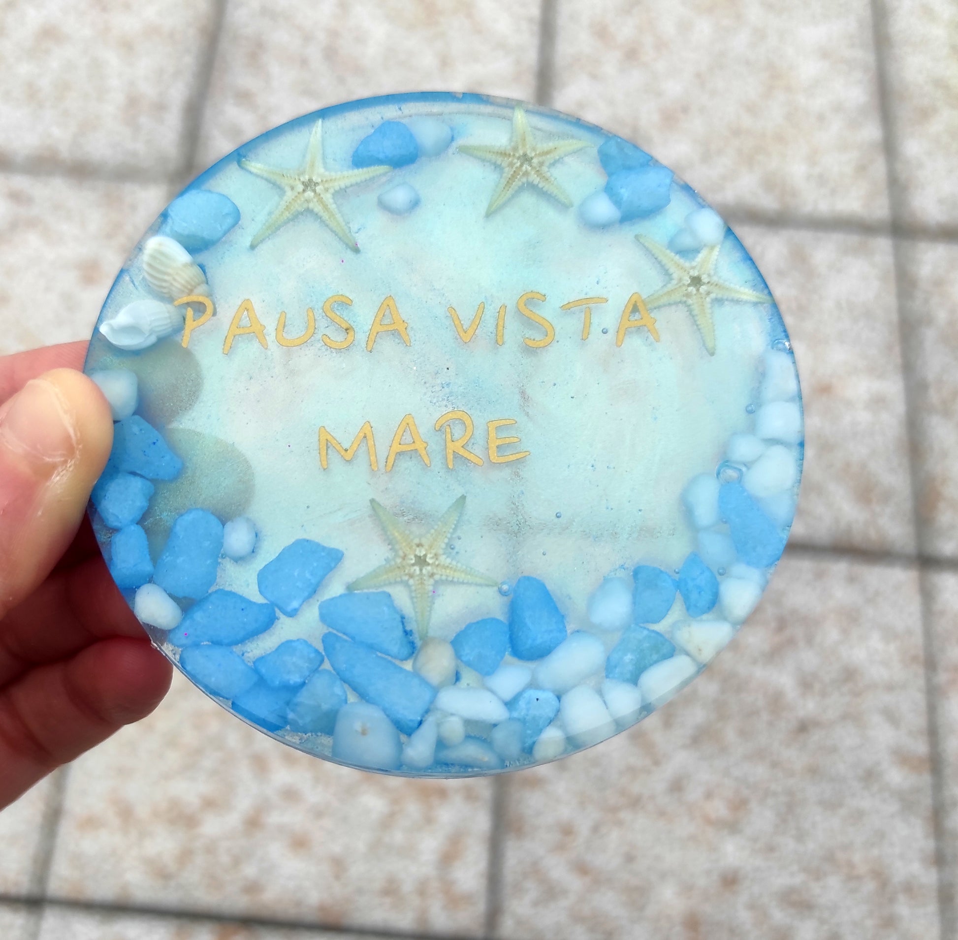 Sottotazza circolare in resina, azzurro, con la scritta dorata "PAUSA VISTA MARE", decorato con stelle marine, conchiglie e pietruzze bianche e celesti.