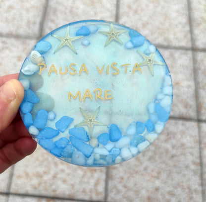Sottotazza circolare in resina, azzurro, con la scritta dorata "PAUSA VISTA MARE", decorato con stelle marine, conchiglie e pietruzze bianche e celesti.