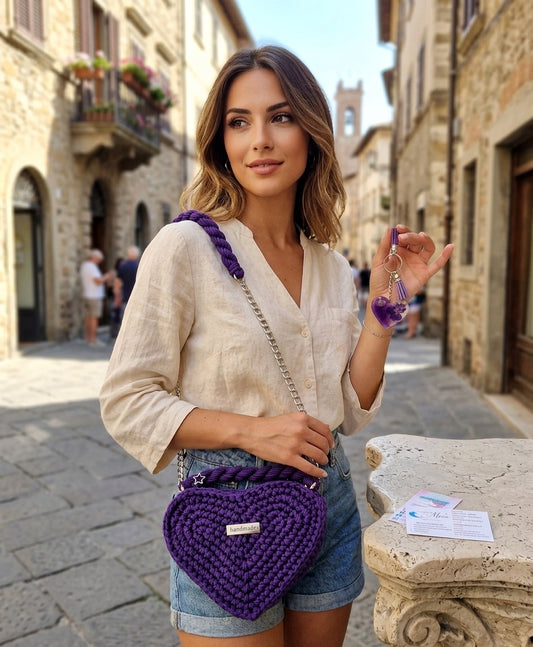 Borsa artigianale all'uncinetto a forma di cuore di colore viola inteso, con manico intrecciato e tracolla in catena metallica argento. Abbinato un portachiavi a forma di cuore in resina viola con fiori secchi e nappa lilla. 