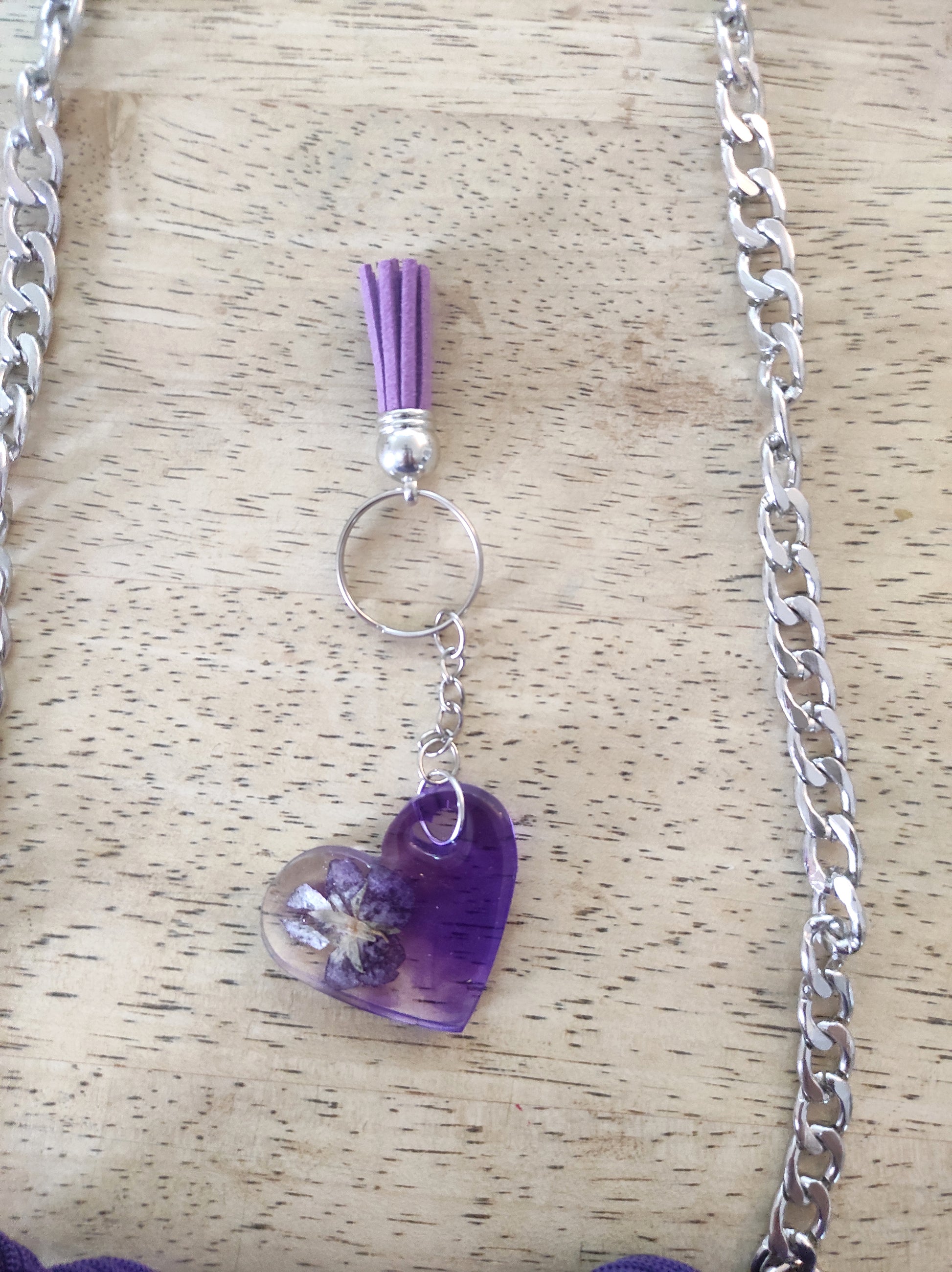 Borsa artigianale all'uncinetto a forma di cuore di colore viola inteso, con manico intrecciato e tracolla in catena metallica argento. Abbinato un portachiavi a forma di cuore in resina viola con fiori secchi e nappa lilla. 