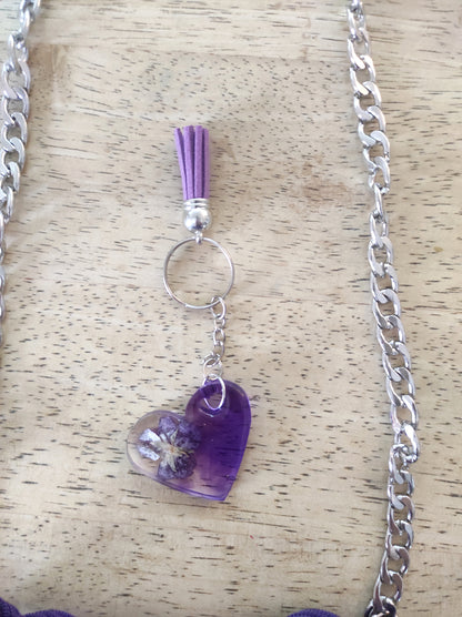 Borsa artigianale all'uncinetto a forma di cuore di colore viola inteso, con manico intrecciato e tracolla in catena metallica argento. Abbinato un portachiavi a forma di cuore in resina viola con fiori secchi e nappa lilla. 
