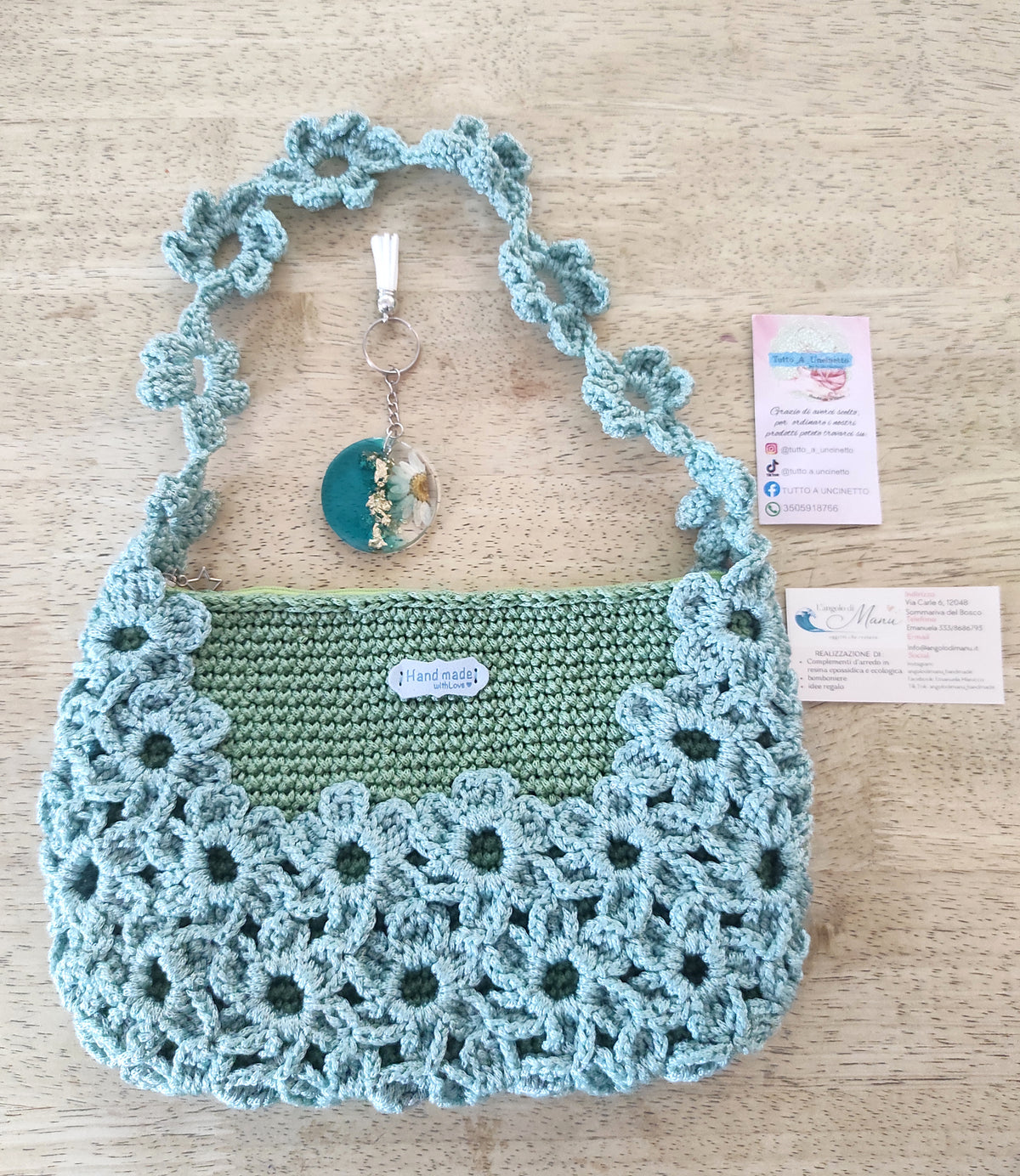 Borsa a mano fatta all'uncinetto nei toni del verde salvia e azzurro polvere, impreziosita da un motivo di fiori intrecciati sulla superficie e lungo il manico. Abbinato un portachiavi rotondo in resina turchese con fiore bianco vero incastonato e dettagli in foglia d'oro