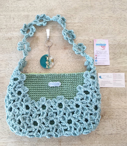 Borsa a mano fatta all'uncinetto nei toni del verde salvia e azzurro polvere, impreziosita da un motivo di fiori intrecciati sulla superficie e lungo il manico. Abbinato un portachiavi rotondo in resina turchese con fiore bianco vero incastonato e dettagli in foglia d'oro
