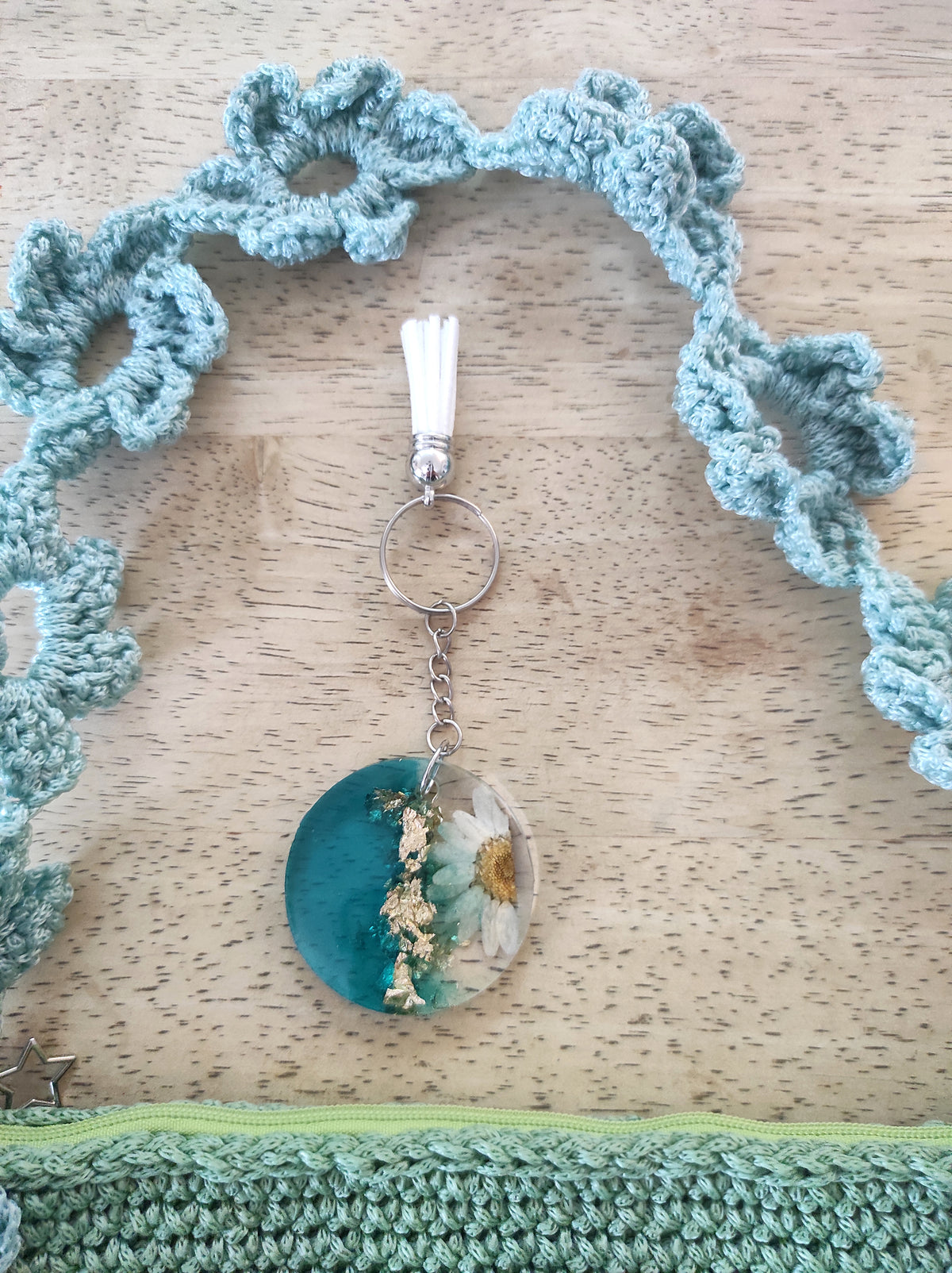Borsa a mano fatta all'uncinetto nei toni del verde salvia e azzurro polvere, impreziosita da un motivo di fiori intrecciati sulla superficie e lungo il manico. Abbinato un portachiavi rotondo in resina turchese con fiore bianco vero incastonato e dettagli in foglia d'oro