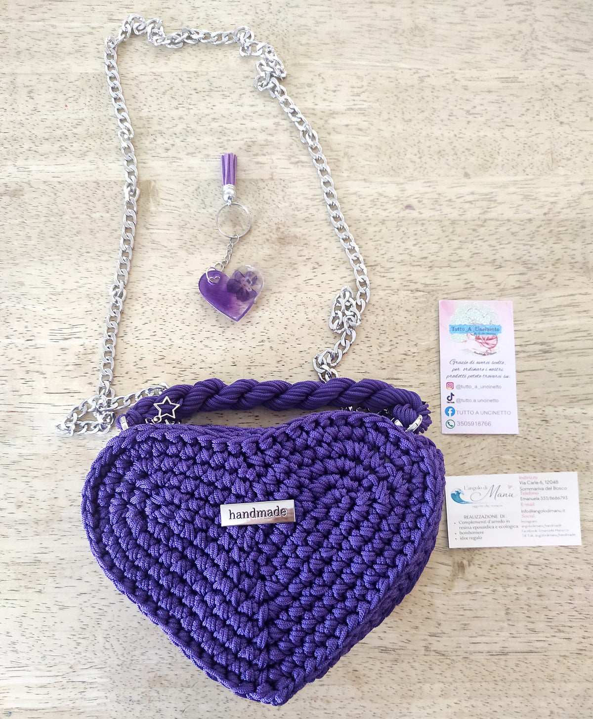 Borsa artigianale all'uncinetto a forma di cuore di colore viola inteso, con manico intrecciato e tracolla in catena metallica argento. Abbinato un portachiavi a forma di cuore in resina viola con fiori secchi e nappa lilla. 