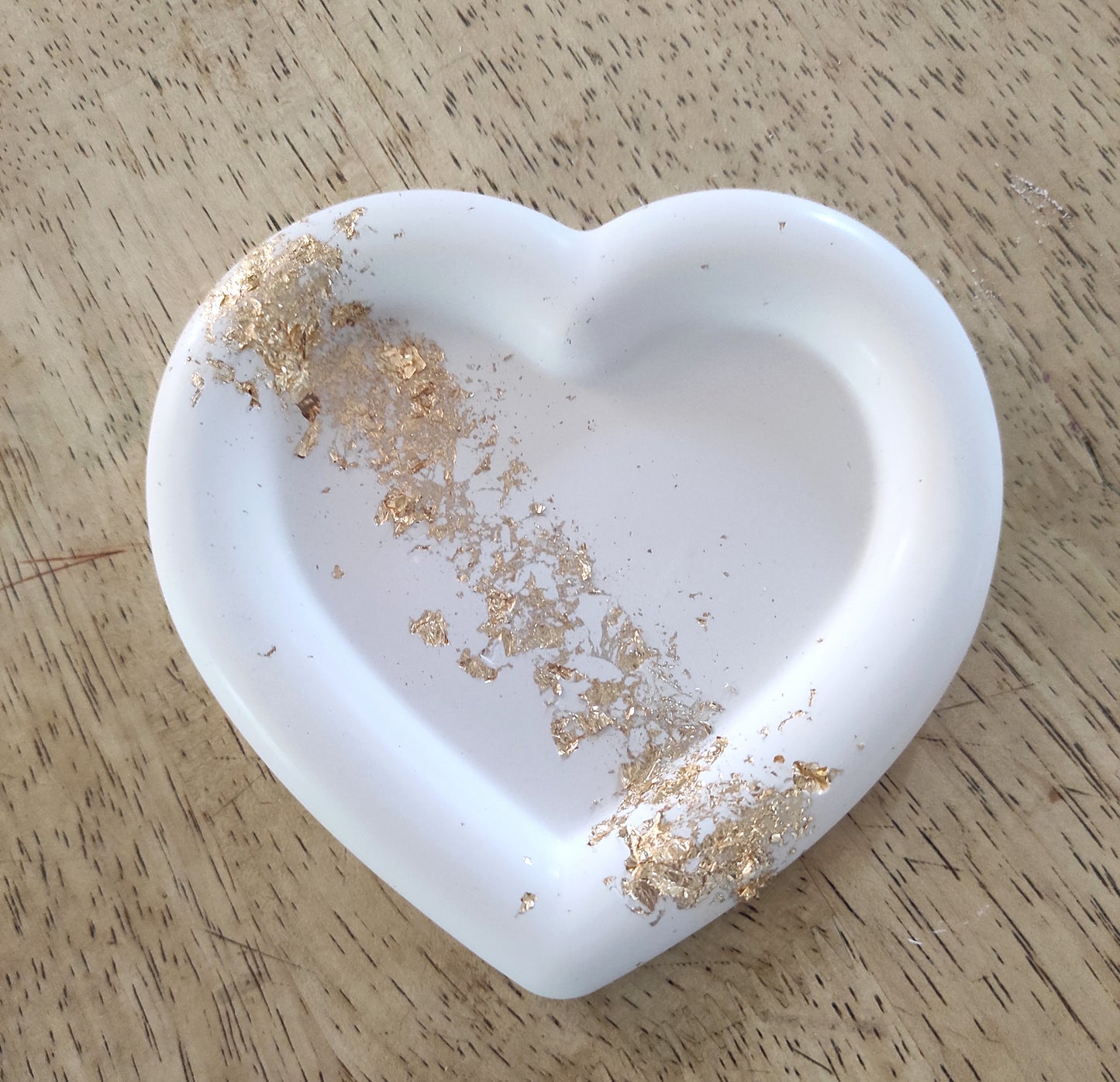 Svuotatasche artigianale a forma di cuore in resina bianca con rifinitura liscia, appoggiato su un piano in legno. L'interno del piatto è decorato con una striscia di dettagli in foglia oro frantumata che attraversa la superficie.