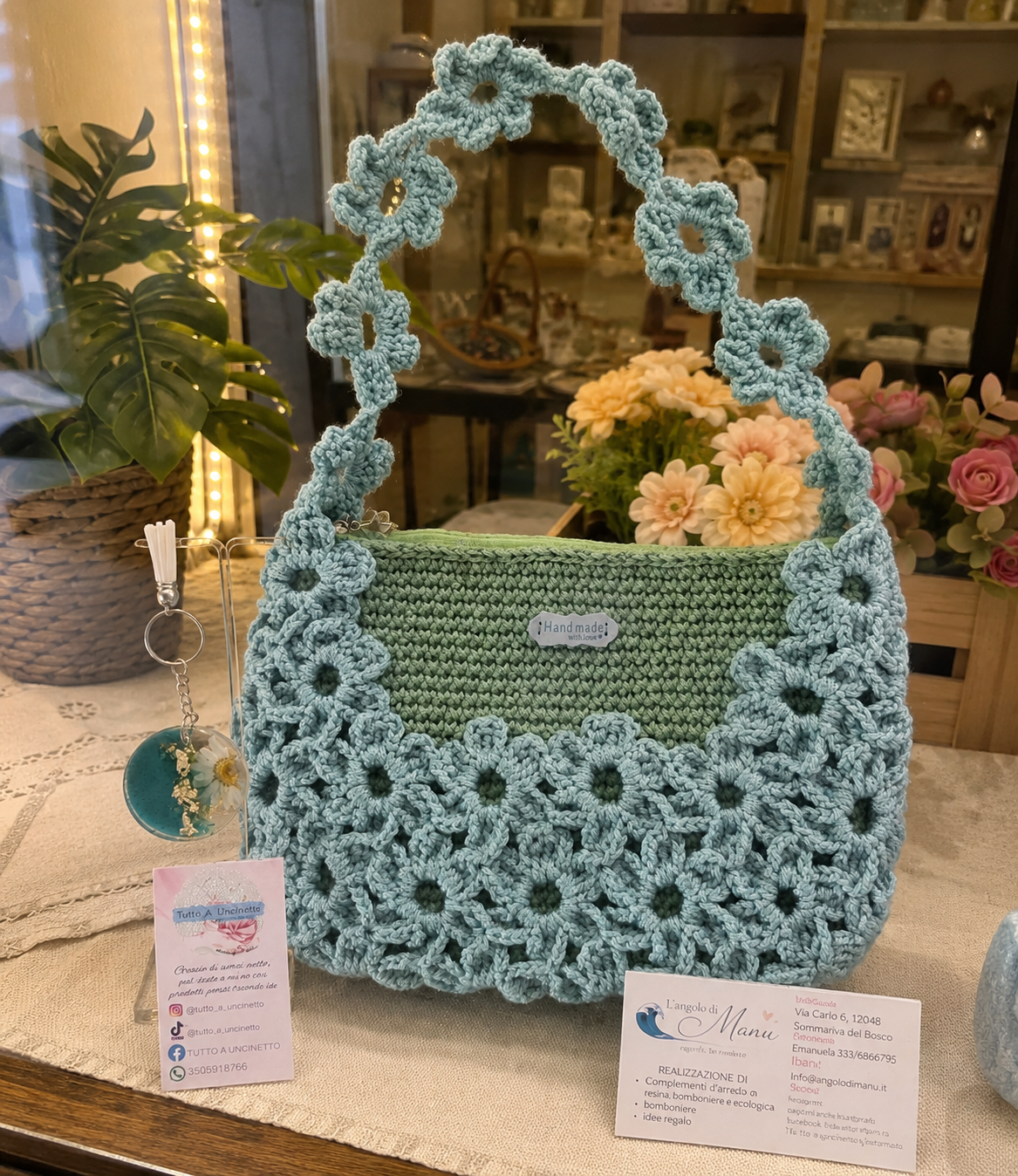 Borsa a mano fatta all'uncinetto nei toni del verde salvia e azzurro polvere, impreziosita da un motivo di fiori intrecciati sulla superficie e lungo il manico. Sulla sinistra, un portachiavi rotondo in resina turchese con fiore bianco vero incastonato e dettagli in foglia d'oro