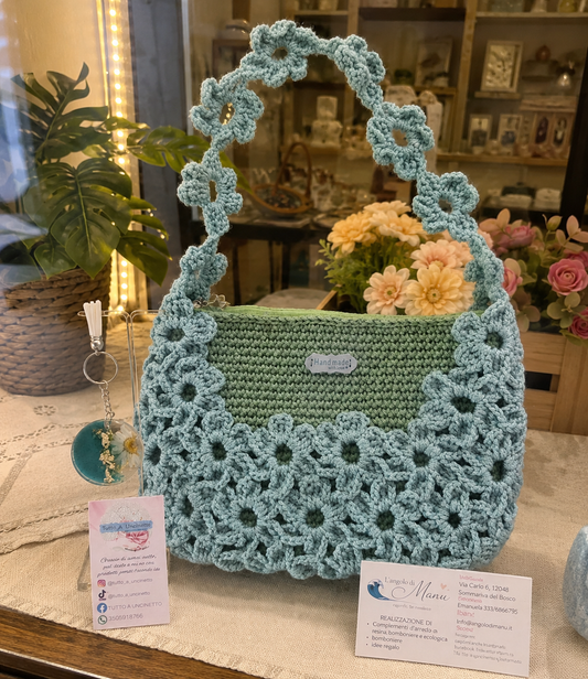 Borsa a mano fatta all'uncinetto nei toni del verde salvia e azzurro polvere, impreziosita da un motivo di fiori intrecciati sulla superficie e lungo il manico. Sulla sinistra, un portachiavi rotondo in resina turchese con fiore bianco vero incastonato e dettagli in foglia d'oro