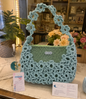 Borsa a mano fatta all'uncinetto nei toni del verde salvia e azzurro polvere, impreziosita da un motivo di fiori intrecciati sulla superficie e lungo il manico. Sulla sinistra, un portachiavi rotondo in resina turchese con fiore bianco vero incastonato e dettagli in foglia d'oro