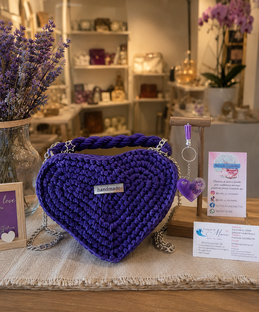 Borsa artigianale all'uncinetto a forma di cuore di colore viola inteso, con manico intrecciato e tracolla in catena metallica argento. Abbinato un portachiavi a forma di cuore in resina viola con fiori secchi e nappa lilla. 