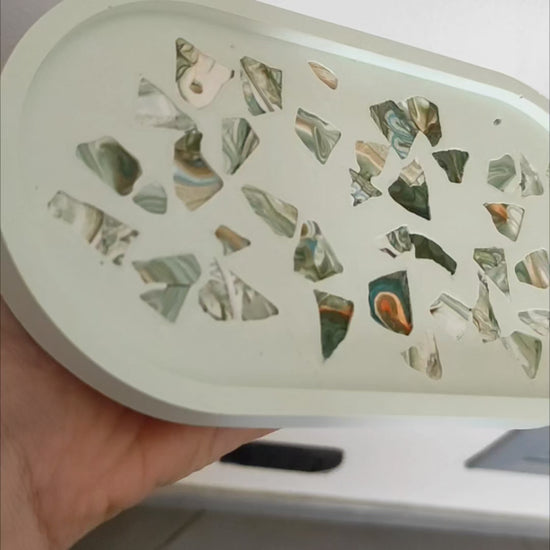 svuotatasche note di verde e marmo: vassoio ovale artigianale in resina ecologica verde pallido. La superficie interna presenta un motivo a terrazzo con frammenti di marmo marmorizzati nei toni del verde scuro, marrone e oro.
