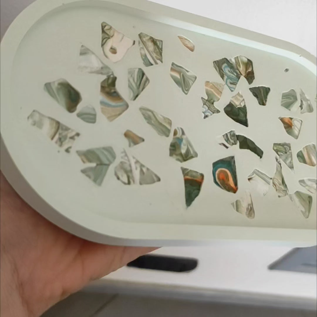 svuotatasche note di verde e marmo: vassoio ovale artigianale in resina ecologica verde pallido. La superficie interna presenta un motivo a terrazzo con frammenti di marmo marmorizzati nei toni del verde scuro, marrone e oro.