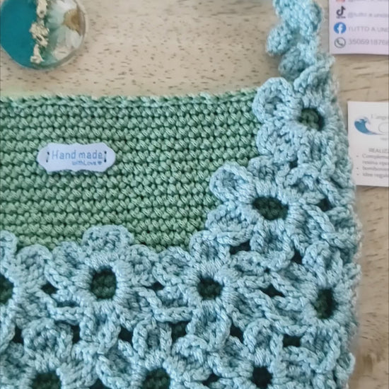Borsa a mano fatta all'uncinetto nei toni del verde salvia e azzurro polvere, impreziosita da un motivo di fiori intrecciati sulla superficie e lungo il manico. Abbinato un portachiavi rotondo in resina turchese con fiore bianco vero incastonato e dettagli in foglia d'oro