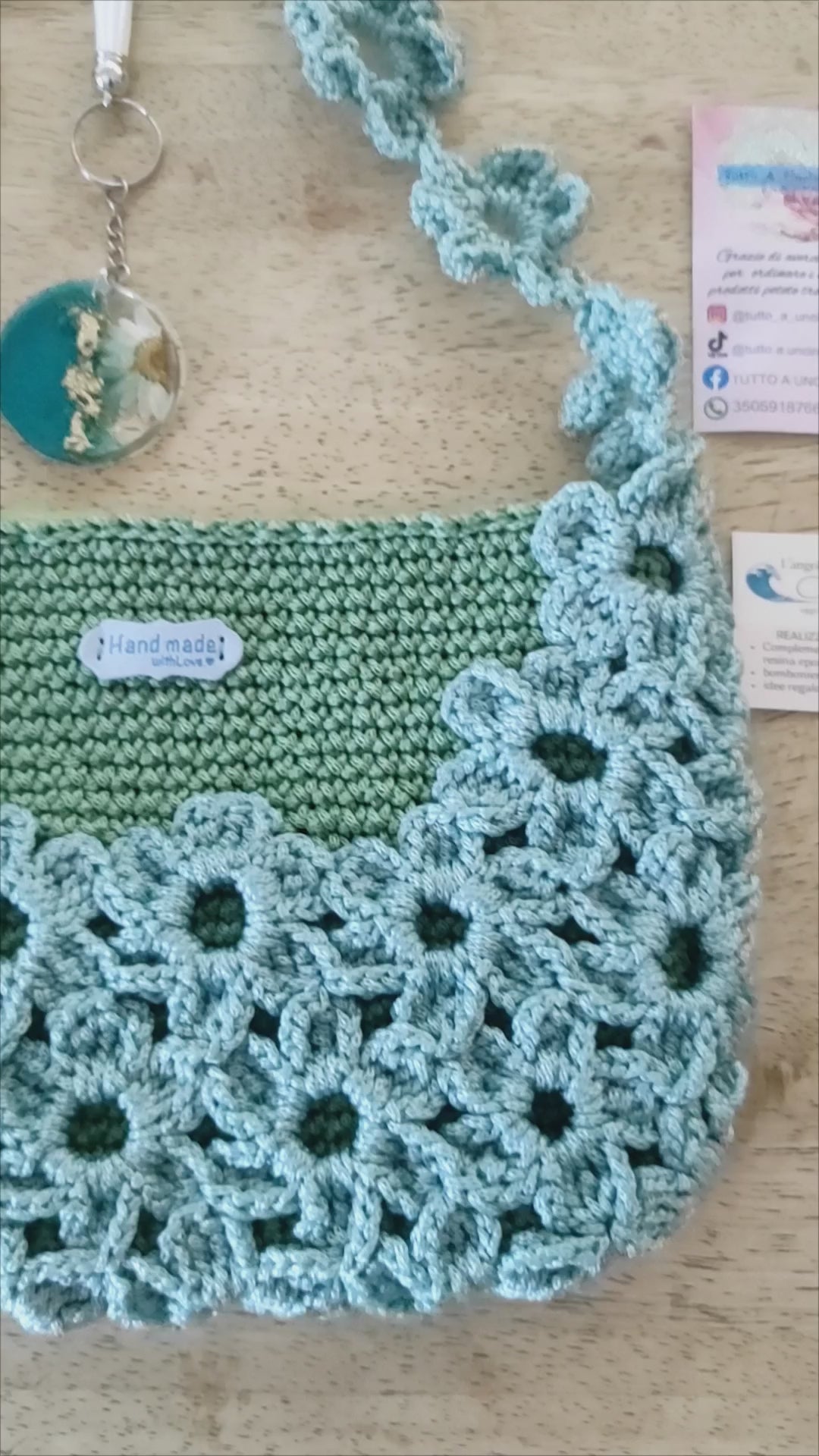 Borsa a mano fatta all'uncinetto nei toni del verde salvia e azzurro polvere, impreziosita da un motivo di fiori intrecciati sulla superficie e lungo il manico. Abbinato un portachiavi rotondo in resina turchese con fiore bianco vero incastonato e dettagli in foglia d'oro